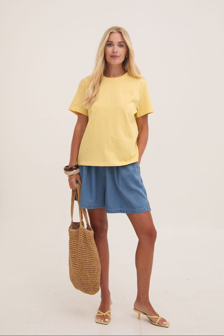 Woman wearing a yellow top and denim shorts holding a woven bag against a white background