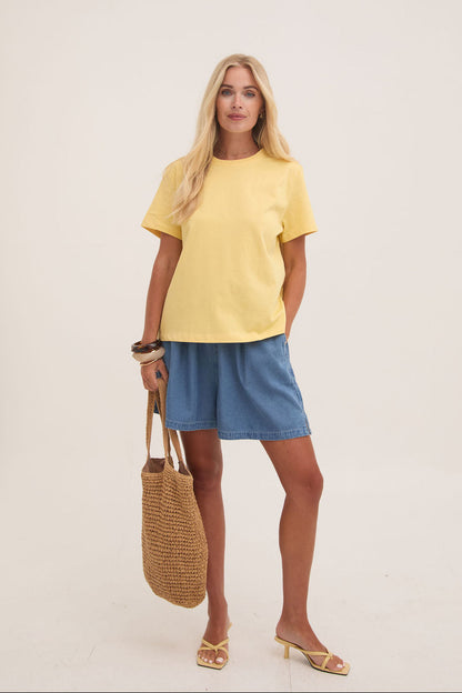 Woman wearing a yellow top and denim shorts holding a woven bag against a white background