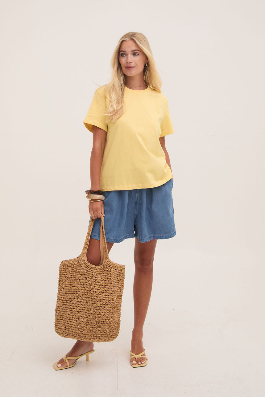 Woman wearing a yellow top and denim shorts, holding a woven bag on a plain background