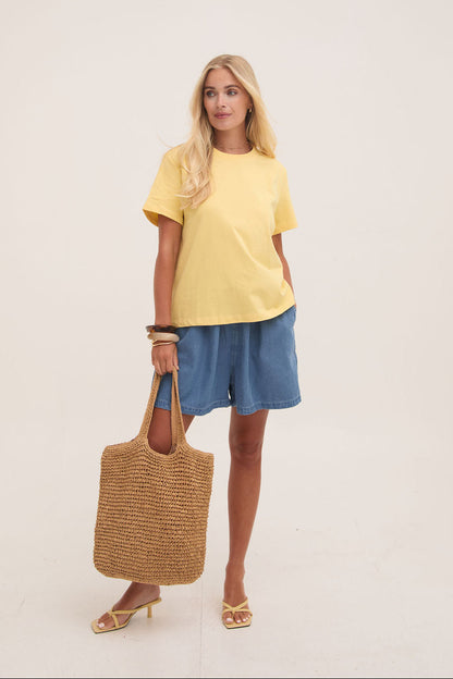 Woman wearing a yellow top and denim shorts, holding a woven bag on a plain background
