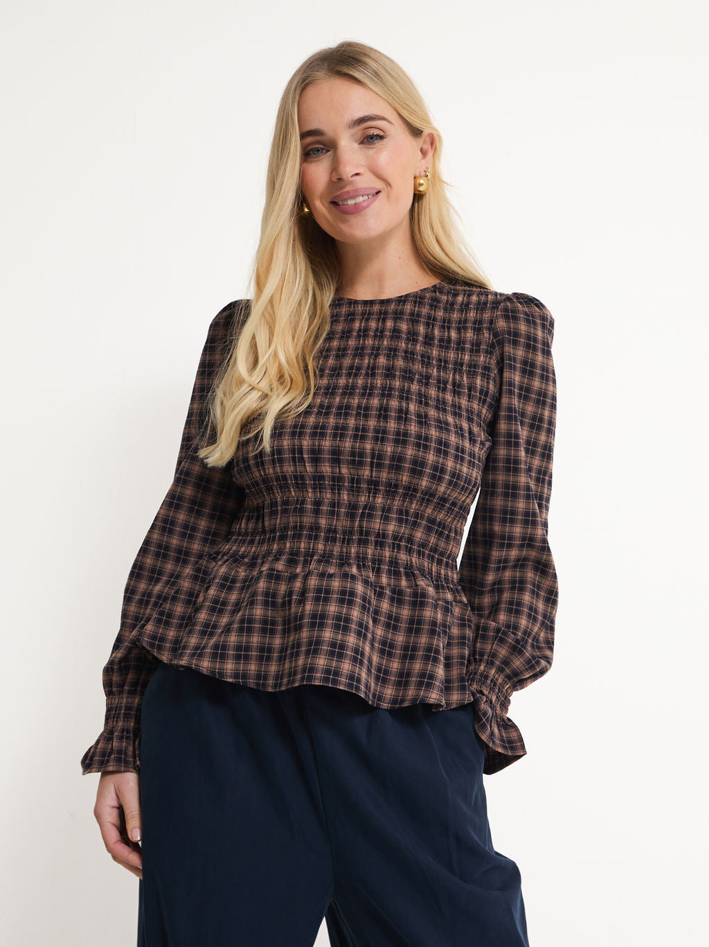 A woman modeling a brown and black plaid long-sleeve blouse with a smocked bodice and peplum hem, paired with dark navy pants, against a plain white studio background.