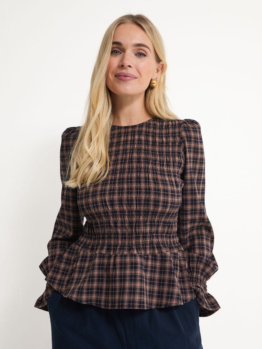 A blonde woman wearing a dark brown and black plaid long-sleeve blouse with a smocked bodice and peplum hem, paired with navy blue pants, against a plain white background.
