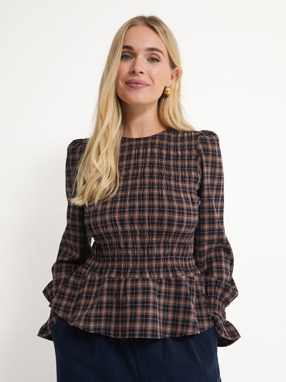A blonde woman wearing a dark brown and black plaid long-sleeve blouse with a smocked bodice and peplum hem, paired with navy blue pants, against a plain white background.