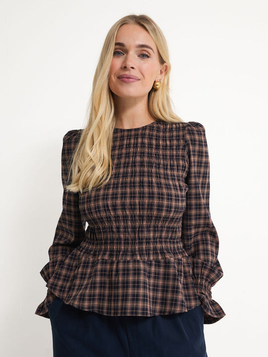 A blonde woman wearing a dark brown and black plaid long-sleeve blouse with a smocked bodice and peplum hem, paired with navy blue pants, against a plain white background.