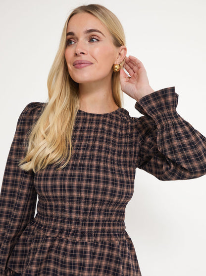 A blonde woman wearing a brown and black plaid long-sleeve dress with a smocked waist, posing against a plain white background.