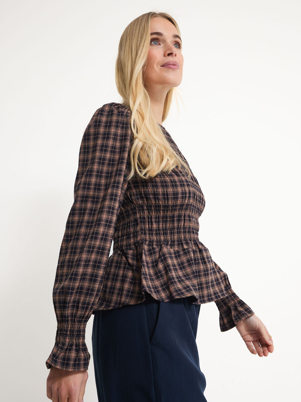 A blonde woman wearing a brown and black plaid long-sleeve blouse with a smocked bodice and peplum hem, paired with navy pants, photographed against a plain white studio background.