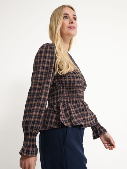 A blonde woman wearing a brown and black plaid long-sleeve blouse with a smocked bodice and peplum hem, paired with navy pants, photographed against a plain white studio background.