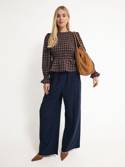 A woman modeling a long-sleeve navy and brown plaid peplum top with wide-leg navy pants, brown pointed shoes, and a brown suede shoulder bag, set against a plain white studio background.