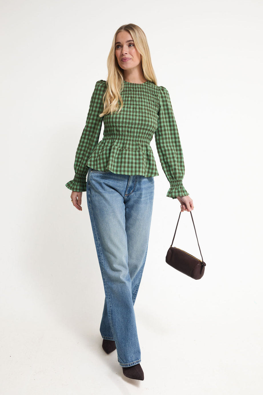A woman modeling a green and black checkered long-sleeve peplum top with ruffled cuffs, paired with light-wash blue jeans and a small dark brown shoulder bag, against a plain white studio background.