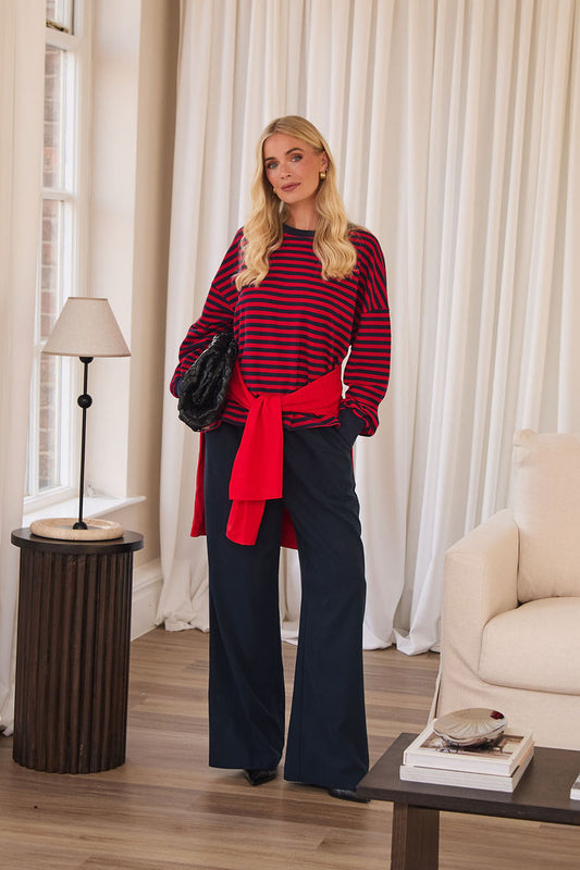 A woman stands in a bright living room wearing a red and navy striped knit sweater with a bright red waist tie, navy wide-leg pants, and holding a black handbag.