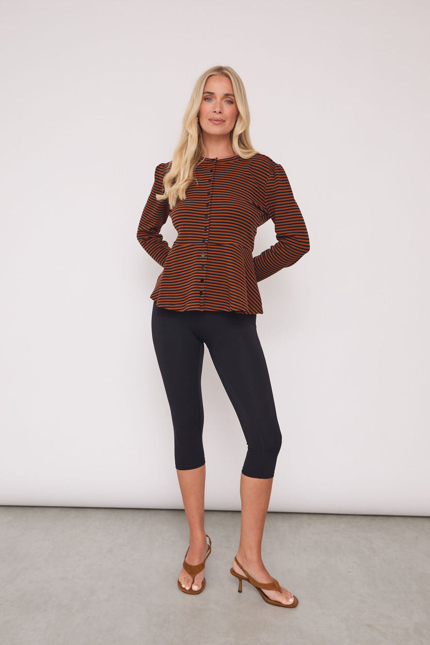 A blonde woman models a brown and black striped long-sleeve button-up top with a peplum waist, paired with black capri leggings and tan heeled sandals, standing against a plain white studio wall.