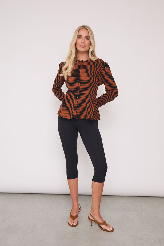 A blonde woman models a brown and black striped long-sleeve button-up top with a peplum waist, paired with black capri leggings and tan heeled sandals, standing against a plain white studio wall.