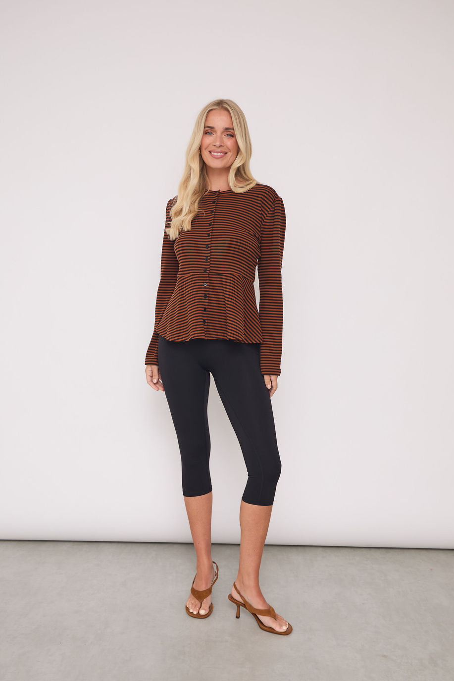 A woman modeling a brown and black striped long-sleeve knit top with a button-up front, paired with black capri leggings and brown heeled sandals, standing against a plain light gray wall.