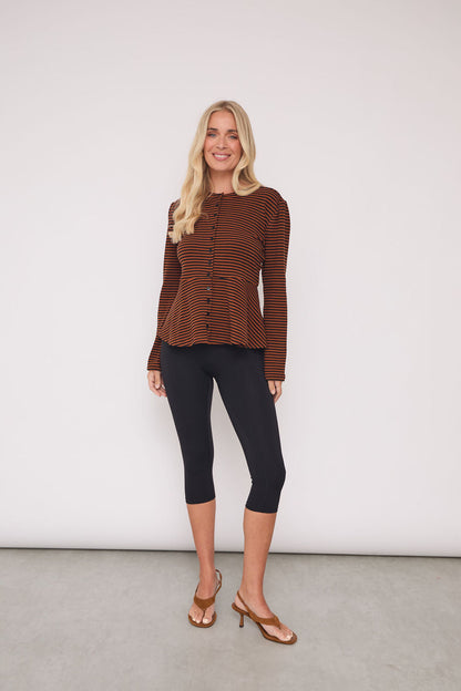A woman modeling a brown and black striped long-sleeve knit top with a button-up front, paired with black capri leggings and brown heeled sandals, standing against a plain light gray wall.
