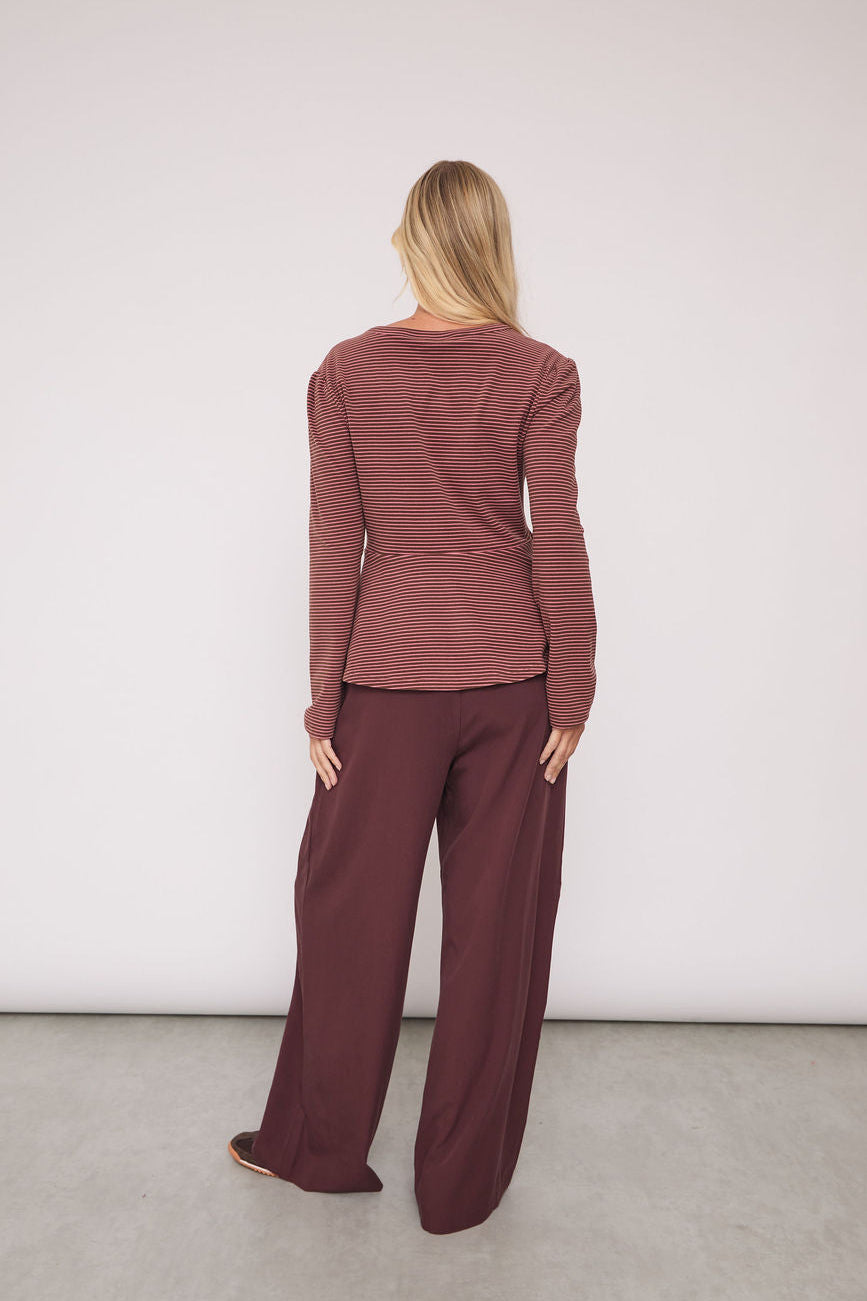 A woman stands with her back to the camera wearing a matching rust-brown long-sleeve top and wide-leg trousers set, photographed in a minimalist studio with a white wall and gray floor.