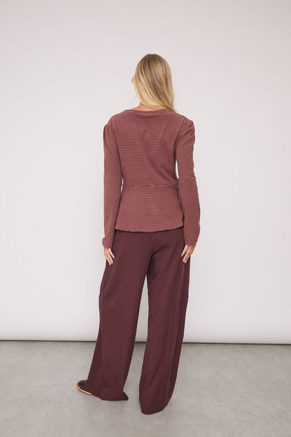 A woman stands with her back to the camera wearing a matching rust-brown long-sleeve top and wide-leg trousers set, photographed in a minimalist studio with a white wall and gray floor.