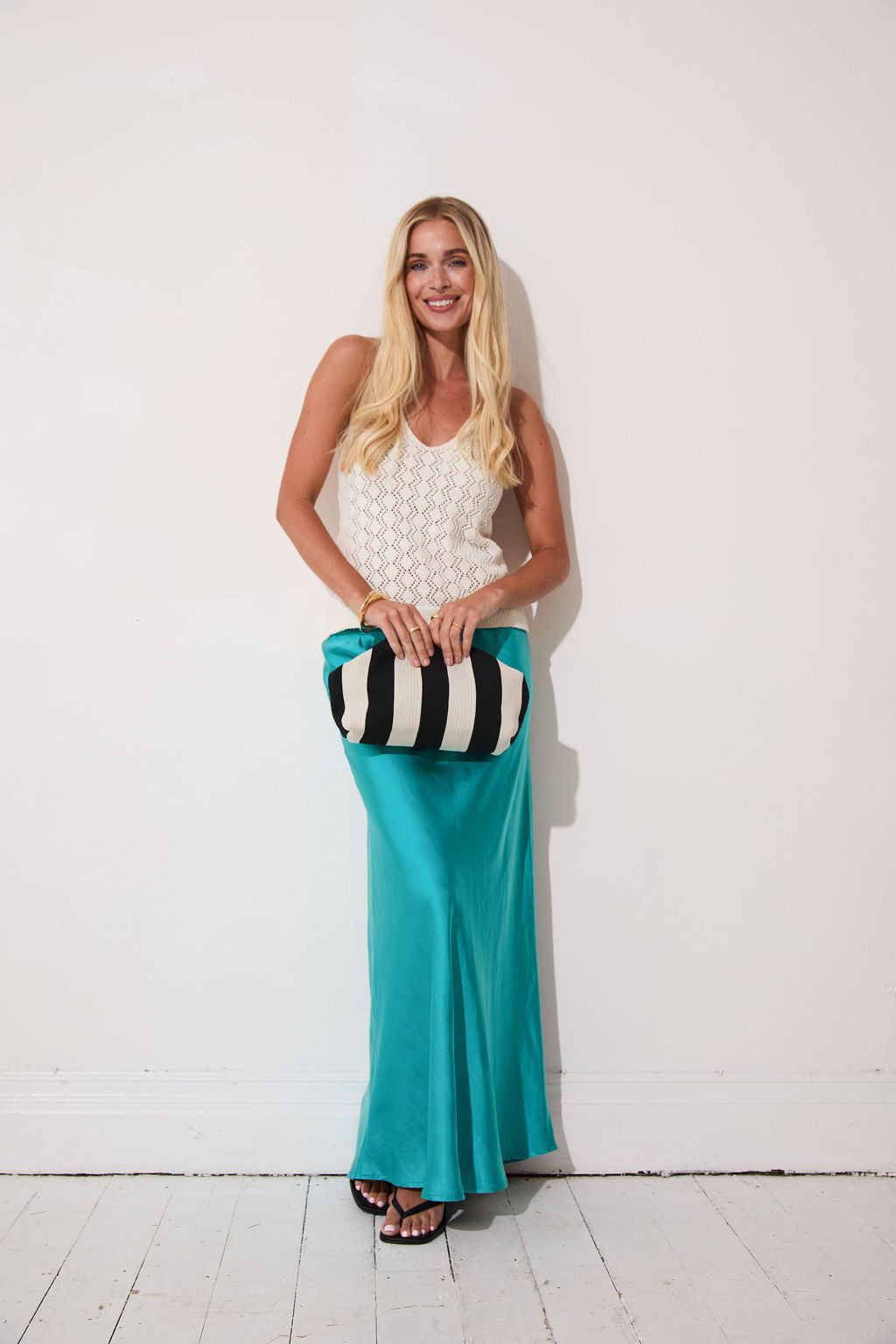 Woman in a white top and turquoise pants holding a striped clutch against a white background