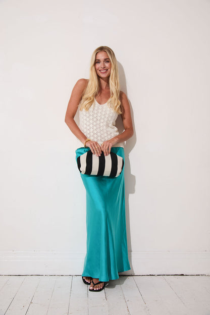 Woman in a white top and turquoise pants holding a striped clutch against a white background